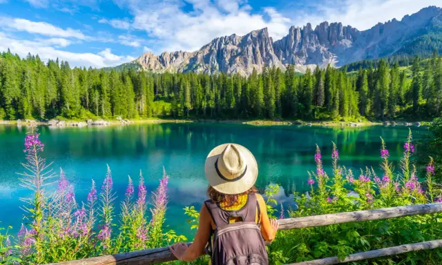 En kvinna med hatt och ryggsäck njuter av den vackra **Dolomiterna sommar**-utsikten över en turkos sjö, omgiven av skog och berg.