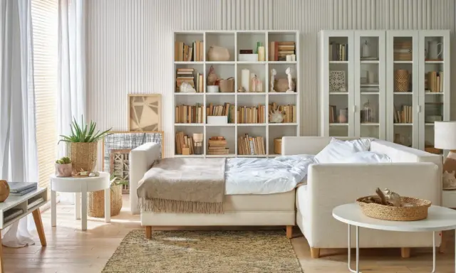 Jasny salon z sypialnią na małej powierzchni. Rozkładana sofa, regały z książkami i dekoracje tworzą przytulną przestrzeń.
