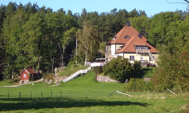 Gula huset vid grinda Stockholm, med röda stugor och grönskande träd.