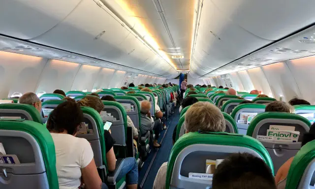 Wnętrze samolotu Transavia, pasażerowie siedzą w rzędach, przygotowani do lotu. Bagaż podręczny schowany w schowkach nad głowami.