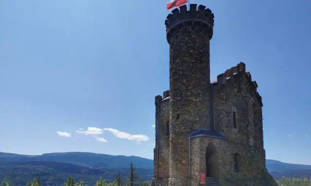 Kamienny zamek henryka na wzgórzu, z polską flagą na wieży, góruje nad zielonymi lasami i błękitnymi górami.