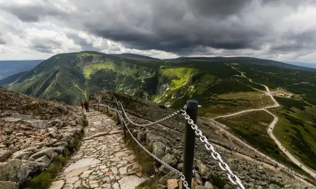 Kamienista ścieżka prowadzi w stronę gór, gdzie widać zielone zbocza i burzowe chmury. Snieżka i jej okolice zachwycają.