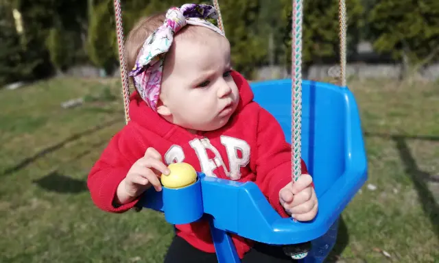 14 miesięczne dziecko w czerwonej bluzie z kapturem i kolorowej opasce na głowie siedzi na niebieskiej huśtawce na zewnątrz.