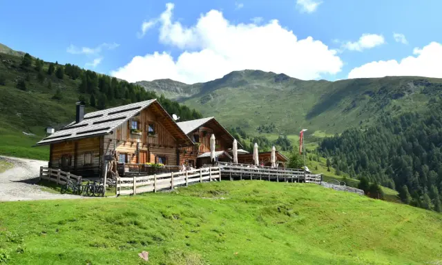 Malga Alto Adige, un rifugio in legno con tavoli all'aperto, circondato da verdi pendii e montagne boscose sotto un cielo azzurro con nuvole bianche.