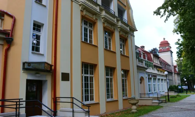 Sanatorium Jan Kazimierz w Dusznikach-Zdroju, z charakterystyczną żółto-kremową fasadą i czerwonym dachem, otoczone zielenią.