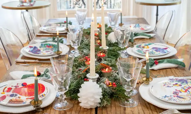 Une table dressée pour les fêtes, avec des assiettes décorées, des bougies allumées et une guirlande de sapin, évoquant la magie des arts de la table.