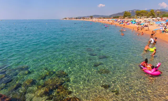 Spiaggia affollata a Santa Susanna, Spagna, con persone che nuotano e si rilassano in un mare cristallino.