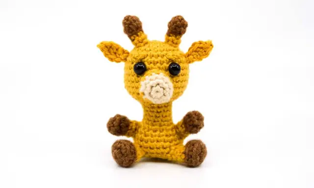Tierna jirafa amigurumi amarilla, sentada con sus patitas marrones. Sus ojos negros y hocico beige le dan un aspecto adorable.