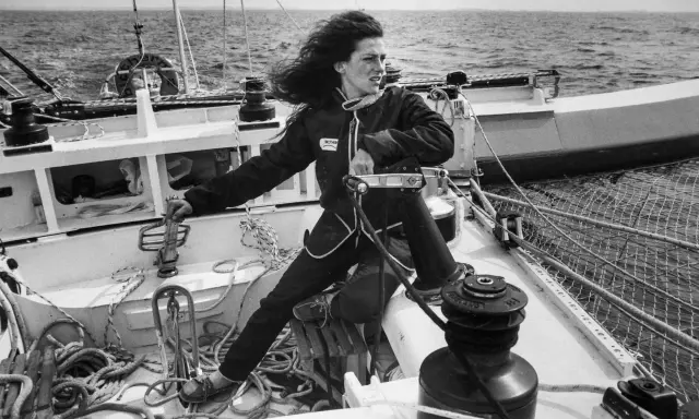 Une jeune femme aux cheveux longs, vêtue d'une combinaison de marin, manœuvre un winch sur un voilier. L'image évoque l'esprit d'aventure de Jacques Arthaud.