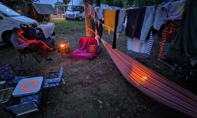 Una persona lee en una silla junto a una fogata, ropa secándose al aire y una hamaca iluminada. Disfrutando de una tranquila acampada al aire libre.