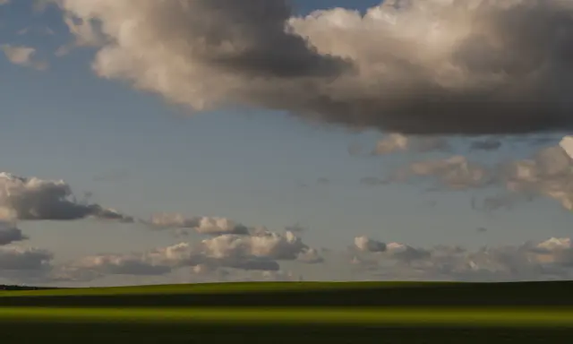 Zachód słońca nad zielonym polem, rzucającym długi cień. Chmury na niebie tworzą ciekawe fotografie.