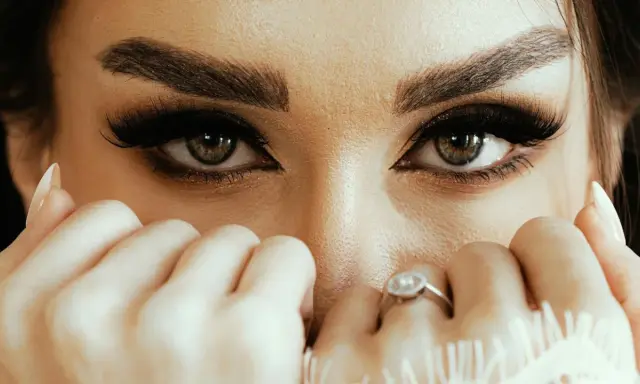 Yeux verts intenses, cils longs et maquillage sourcils parfait. Mains délicates ornées de bagues cachent une partie du visage.