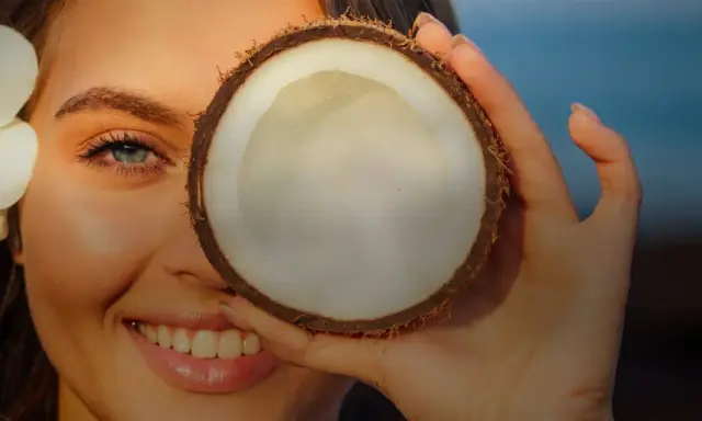Donna sorride con un fiore tra i capelli, coprendo un occhio con una noce di cocco. Attenzione: olio di cocco sul viso fa male.