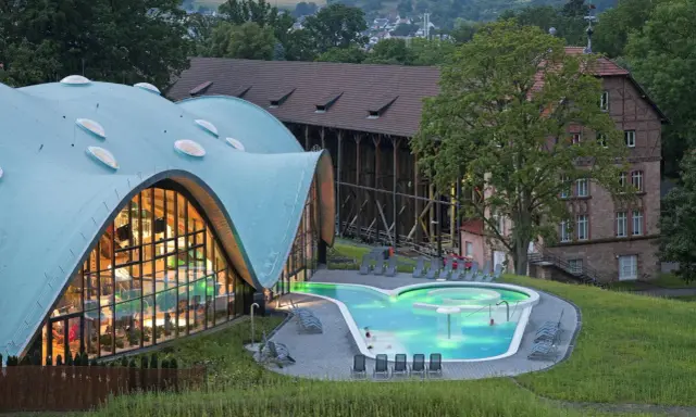 Moderne Therme mit geschwungenem Dach und beleuchtetem Pool, daneben historische Gebäude.