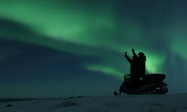 En person på snöskoter i Kiruna Lapland sträcker upp armarna mot norrskenet som lyser grönt på natthimlen.