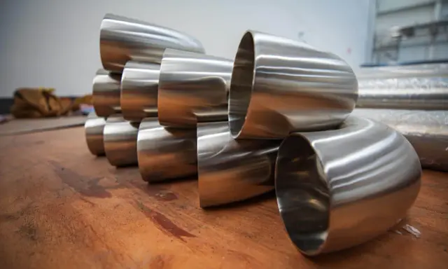 Pièces métalliques brillantes, probablement des coudes pour un poêle à bois conduit, empilées sur une surface en bois.