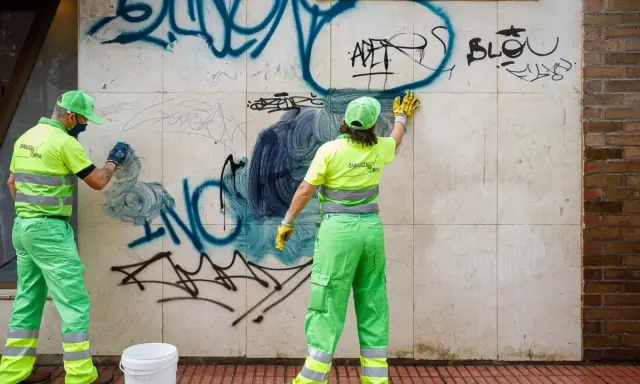 Dos trabajadores con uniformes verdes y amarillos brillantes trabajan para limpiar grafitis de una pared de mármol.