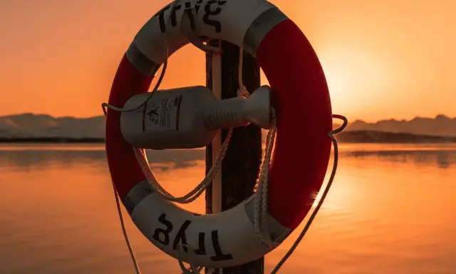 Bouée de sauvetage rouge et blanche sur un poteau, au bord d'une voie d'eau calme, avec un bateau à l'horizon au coucher du soleil.