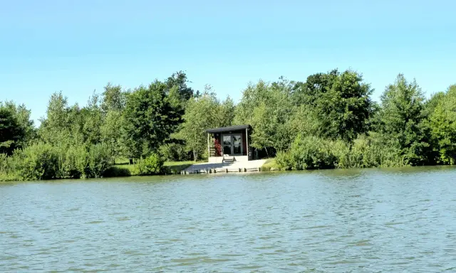 Spokojne wody magnum carp lake otaczają zielone drzewa. Mały domek z tarasem stoi przy brzegu.