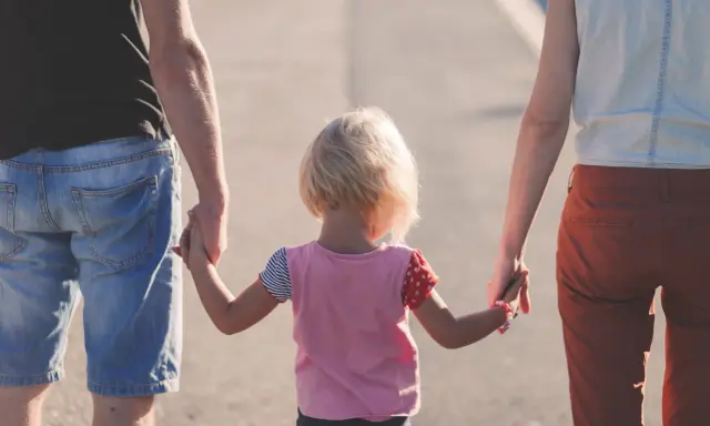 Rodzina idzie razem, trzymając się za ręce. Dziecko z blond włosami, w kolorowej bluzce, między rodzicami. Może to być obraz rodziny, która radzi sobie z zaburzeniami zachowania ICD 10.