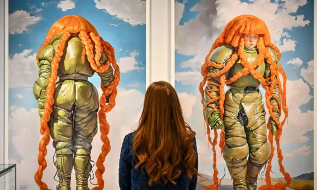 A woman admires two surreal paintings of figures in puffy green suits with bright orange braided hair at Christie's NYC.