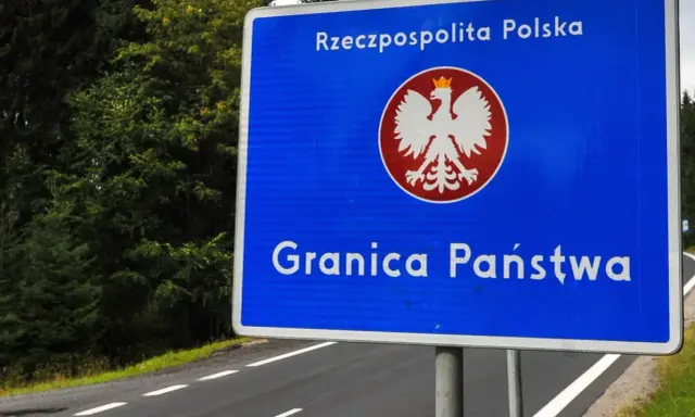 Niebieski znak z orłem i napisem "Rzeczpospolita Polska" oraz "Granica Państwa" stoi przy drodze otoczonej lasem.