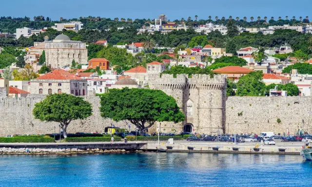 Widok na zabytkowe mury obronne i zabudowania wyspy Rodos, z błękitnym morzem w tle.