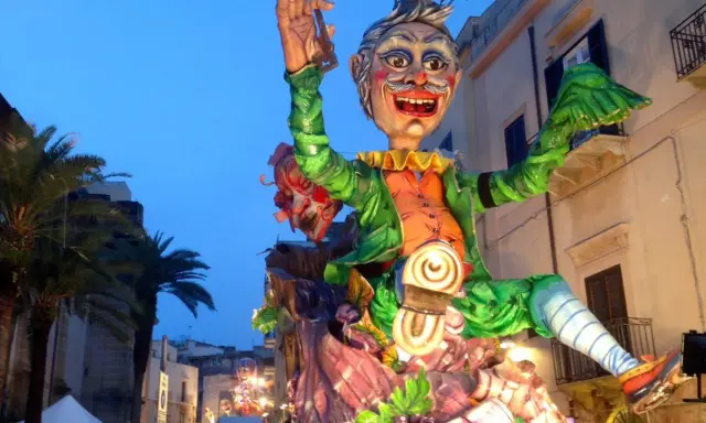 Carro allegorico con maschere di carnevale siciliane, un personaggio gioioso con vestiti verdi e arancioni sfila tra palme e palazzi illuminati.