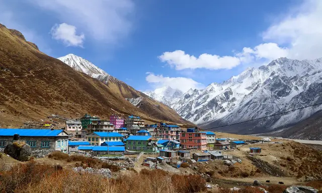 Villaggio colorato ai piedi dell'alta montagna himalayana, con case dai tetti blu e rosa, circondato da pendii bruni e cime innevate.