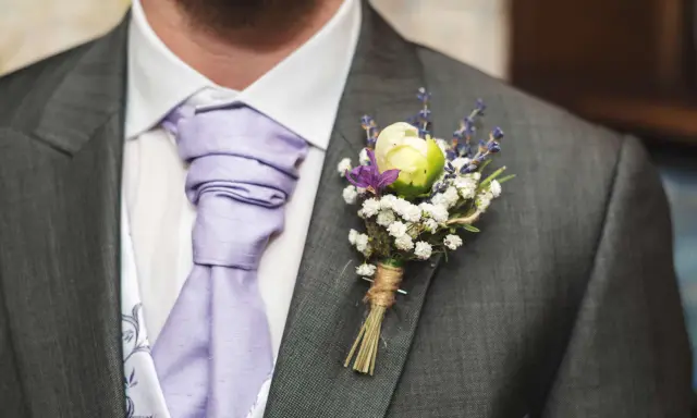Elegancka butonierka w marynarce, ozdobiona białymi kwiatkami i lawendą, dodaje uroku ślubnemu strojowi.