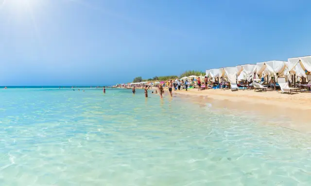 Spiaggia affollata con lettini e ombrelloni bianchi, gente che nuota e prende il sole. Un'immagine perfetta delle località balneari Puglia.