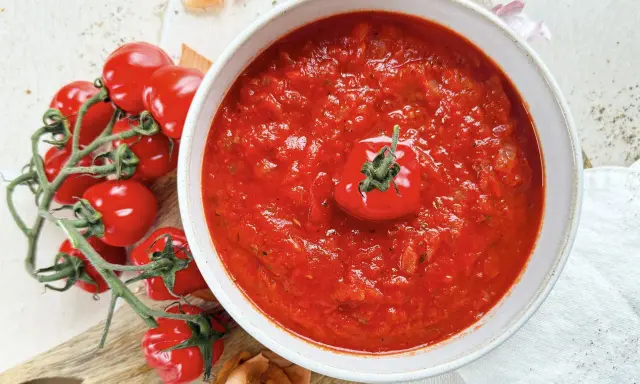 Eine Schüssel mit köstlicher, hausgemachter italienischer Tomatensauce, garniert mit einer frischen Kirschtomate. Daneben liegen frische Tomaten und Zwiebeln.