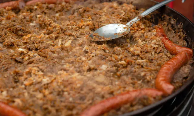 Wielki gar bigosu z kiełbasą, gotowy do wekowania w piekarniku. Łyżka zanurzona w aromatycznym daniu.