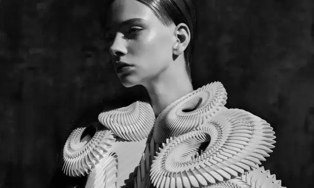 A model showcases intricate, sculptural white garments, embodying futuristic fashion. The avant-garde design features layered, organic shapes that create a dramatic silhouette.