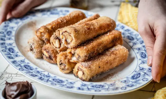 Nutella French Toast Rolls, goldbraun gebacken und mit Zimt-Zucker bestreut, auf einem Teller mit blauem Blumenmuster.
