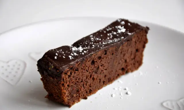Wilgotny brownie z czerwonej fasoli, polany czekoladą i posypany cukrem pudrem, na białym talerzu z serduszkami.