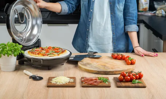 Otwarta opiekacz do pizzy, na kamieniu piec pizza z pomidorkami i bazylią. Obok deska z pomidorkami, startym serem i szynką.