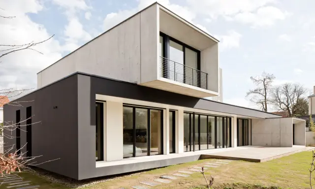 Une maison moderne aux lignes épurées, avec des blocs de béton et de crépi sombre, dotée de grandes baies vitrées et d'une terrasse en bois.
