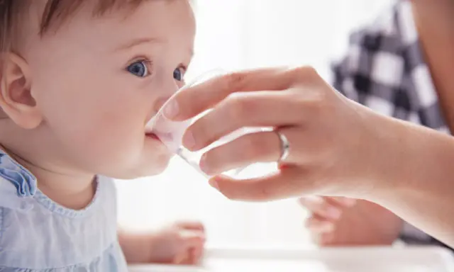 Ein Baby trinkt vorsichtig Wasser aus einem Becher, gehalten von einer Hand. Die Frage "ab wann Baby Wasser geben?" wird hier visuell beantwortet.