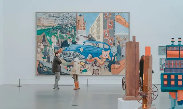 Two people observe a large, colorful mural depicting a chaotic scene with a blue car, factories, and people, offering a glimpse into the art market definition of diverse artistic expression.
