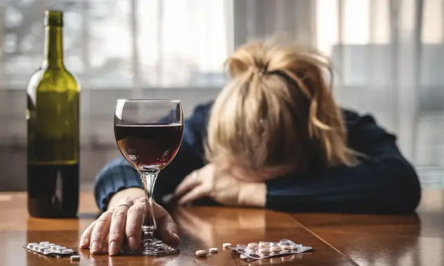 Kobieta z głową opartą na rękach, obok kieliszek wina, butelka i opakowania psychotropów.