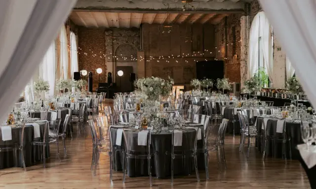 Eleganckie wesele w industrialnym stylu. Stoły nakryte szarymi obrusami, ozdobione białymi kwiatami i transparentnymi krzesłami. Girlandy świateł tworzą magiczny nastrój.