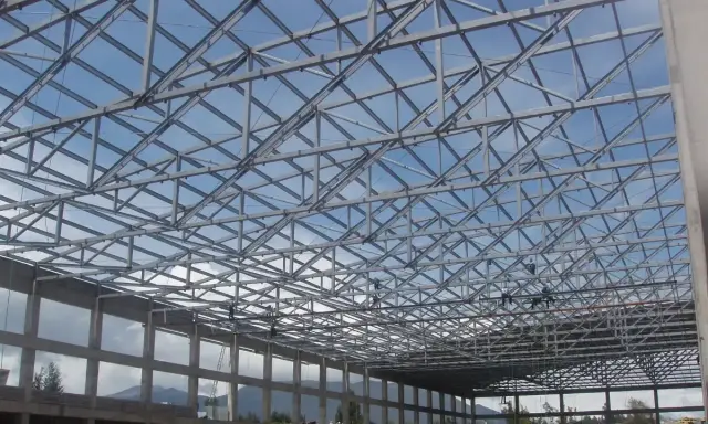Estructura metálica de una **cercha Pratt** en construcción, con operarios trabajando en el techo bajo un cielo azul.