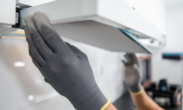 Hände mit Handschuhen ziehen einen Filter aus einer Dunstabzugshaube, um den Motor zu reinigen.
