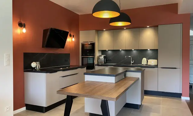 Cuisine moderne aux murs terracotta, avec îlot central en bois et mobilier gris clair. Éclairage chaleureux.