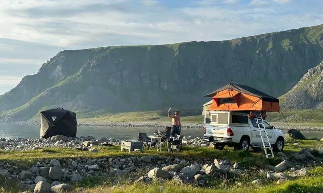 En man tar en selfie vid sitt tak-tält på en strand i Norge. Bredvid står en mörk kupoltält och ett bord med stolar. Berg i bakgrunden.