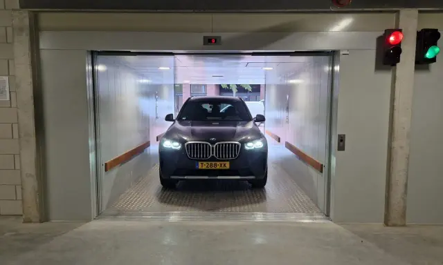 Un coche BMW oscuro espera en un montacoches para garaje. La luz roja del semáforo indica que debe esperar.