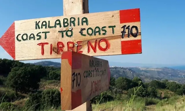 Cartello in legno indica "Kalabria Coast to Coast" verso il Tirreno e lo Jonio, con sfondo di colline e mare.