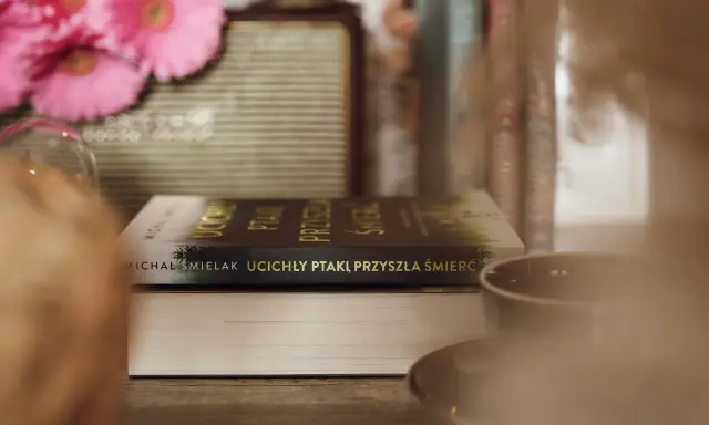 Na stole leżą książki, jedna z nich ma tytuł "Ucichły ptaki, przyszła śmierć". W tle różowe kwiaty i filiżanka.