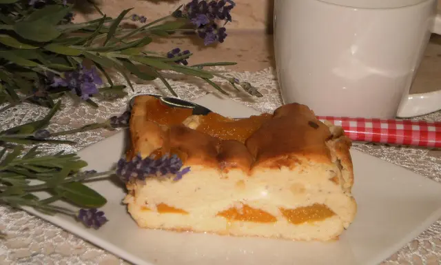 Pyszny sernik z brzoskwiniami, udekorowany gałązką lawendy, obok białej filiżanki i czerwono-białej łyżeczki.